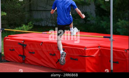 Rückansicht eines jungen männlichen Athleten, der über eine gelbe Hochsprungstange auf rote Matten springt. Stockfoto