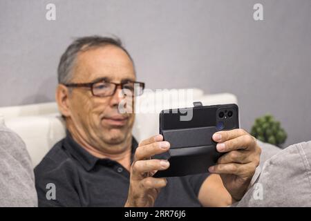 Ein älterer Mann mit Brille, der auf der Couch sitzt, verwendet ein Smartphone Stockfoto