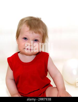 Ein einjähriges Kleinkind mit blondem Haar im roten Body, das auf weißem Hintergrund mit aufblasbaren Ballons sitzt und lächelt. Wunderbares Kind, das sich hinsetzte, um sich auszuruhen Stockfoto