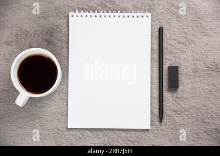 Kaffeetasse mit erhöhter Aussicht, leerer spiralförmiger Notizblock mit schwarzem Radiergummi, grauer Schreibtisch Stockfoto