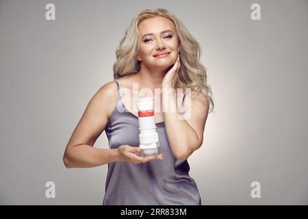 Lächelnde ältere Frau, die verschiedene Gläser Creme über grau gehalten hat Stockfoto