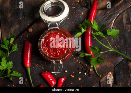 Würzige hausgemachte Chilisauce in einem kleinen Glas auf einem dunklen Holzbrett, mit roten heißen Paprika und Petersilie Stockfoto