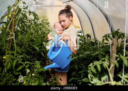 Frau in ihren 20ern mit Baby in den Armen tränkt Tomatensämlinge im Gewächshaus aus der Gartengießkanne. Stockfoto