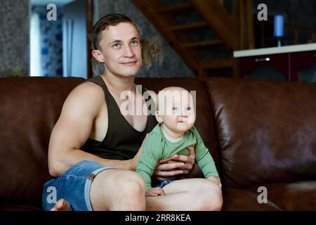 Der junge Vater mit dem sechs Monate alten Säugling sitzt auf dem Ledersofa im Wohnzimmer. Stockfoto