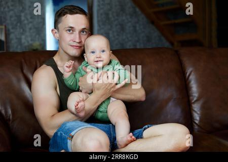 Junger Mann Vater Vater Vater sitzt auf Ledersofa mit seinem sechs Monate alten Baby. Stockfoto