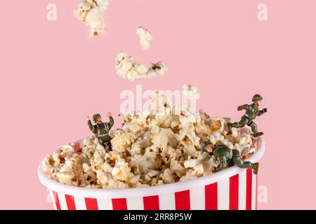 Popcorn in rot-weißem Eimer auf rosa mit kleinen Spielzeugsoldaten, die springen Stockfoto