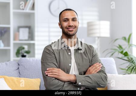 Porträt eines jungen muslimischen Mannes, der zu Hause auf der Couch sitzt und lächelnd die Kamera anschaut. Stockfoto