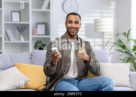 Porträt eines jungen afroamerikanischen Mannes, der zu Hause auf dem Sofa sitzt und die Fernbedienung der Klimaanlage hält, die Kamera anlächelt und die Supergeste mit dem Finger zeigt. Stockfoto