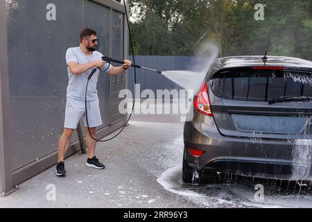 Der Mann hat eine Wasserkanone in der Hand, mit der sie das Auto wäscht. Ein Auto in einer Selbstbedienungswaschanlage. Stockfoto
