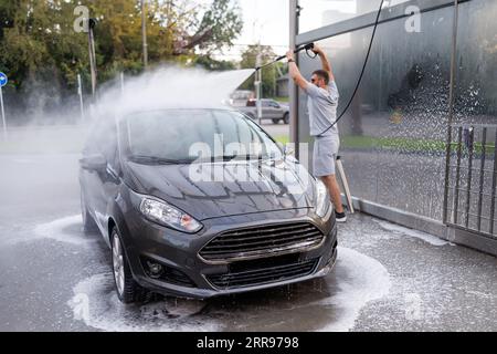 Der Mann im Hintergrund hat die Wasserkanone über seinen Kopf gehoben und zielt auf das Dach des Autos. Ein Auto in einer Selbstbedienungswaschanlage. Stockfoto