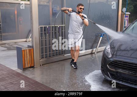 Ein Mann mit einer Wasserkanone in der Hand läuft um das Auto herum und wäscht es. Ein Auto in einer Selbstbedienungswaschanlage. Stockfoto