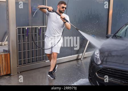 Ein Mann mit einer Wasserkanone in der Hand läuft um das Auto herum und wäscht es. Ein Auto in einer Selbstbedienungswaschanlage. Stockfoto