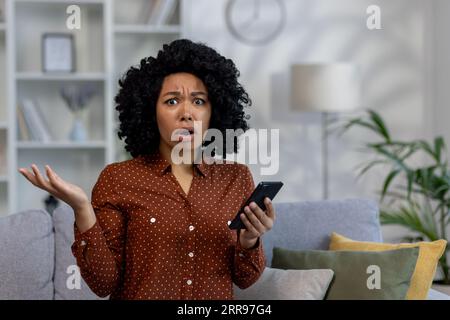 Porträt einer afroamerikanischen Frau, die überrascht und enttäuscht von der Kamera aussieht, auf der Couch sitzt, das Telefon hält und ihre Hände spreizt. Stockfoto