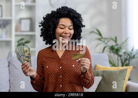 Lächelnde afroamerikanische Frau, die Bargeld in der Hand hält und auf Kreditkarte schaut. Zu Hause auf der Couch sitzen. Stockfoto