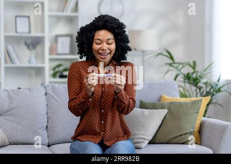 Afroamerikanische junge Frau, die einen Einweg-Schwangerschaftstest zu Hause in den Händen hält, es betrachtet und mit einem positiven Ergebnis zufrieden ist. Stockfoto