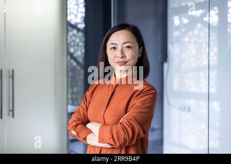 Porträt einer jungen, schönen, selbstbewussten asiatischen Geschäftsfrau, die in einem Bürozentrum steht, mit gekreuzten Armen auf der Brust und ernsthaft auf die Kamera schaut. Stockfoto