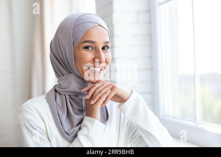 Das Foto ist ein Nahaufnahme-Porträt einer jungen, schönen muslimischen Frau in einem Hijab, die innen am Fenster steht, ihr Gesicht auf ihre Hände lehnt und lächelnd für die Kamera posiert. Stockfoto