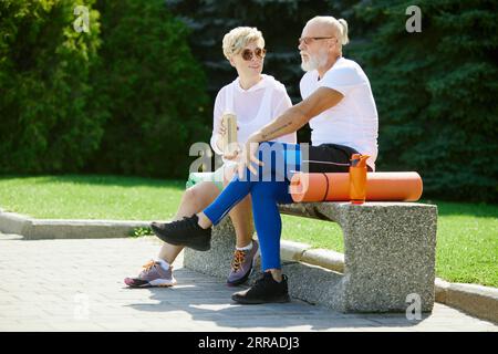 Fröhliche, positive, lächelnde reife Menschen, Mann und Frau, die auf einer Bank im Freien mit Fitnessmatte sitzen. Nach dem Training Stockfoto