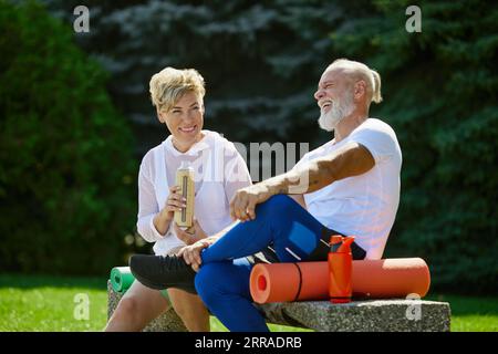Fröhliche, positive, lächelnde reife Menschen, Mann und Frau, die auf einer Bank im Freien mit Fitnessmatte sitzen. Nach dem Training Stockfoto