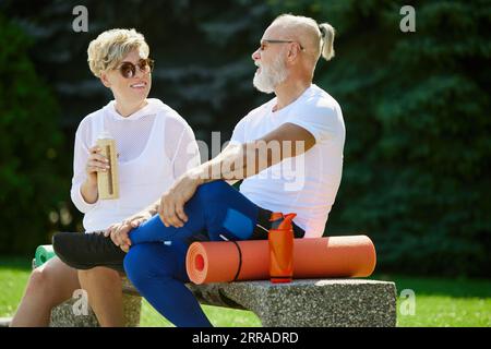 Fröhliche, positive, lächelnde reife Menschen, Mann und Frau, die auf einer Bank im Freien mit Fitnessmatte sitzen. Nach dem Training Stockfoto