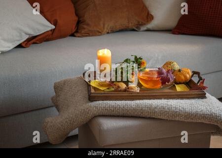 Tasse heißen Tee auf einem Holztablett mit Kerze, Blumen, Kürbissen und Keksen auf einer Wolldecke auf der Couch, gemütlicher Herbst zu Hause, Kopierraum, Stockfoto