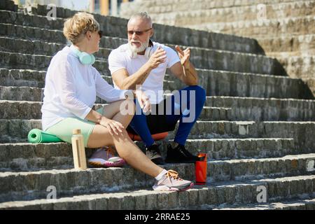 Positive, lächelnde reife Menschen, Mann und Frau, die draußen auf einer Bank sitzen und reden. Nach dem Training Stockfoto