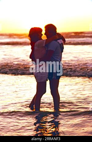 Ein Paar, das während des Sonnenaufgangs am Strand steht und sich umgibt. Stockfoto