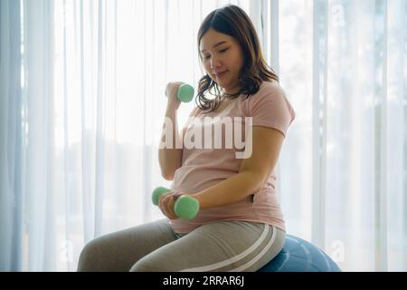 Strahlende asiatische Mom-to-be umarmt Wellness und formt Kraft in ihrem Heim Heiligtum. Kurzhanteln werden zu Werkzeugen der Ermächtigung auf ihrer anmutigen Reise. Stockfoto