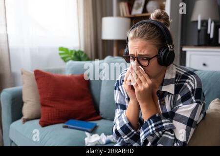 Junge traurige Frau, die allein zu Hause auf dem Sofa sitzt und traurige Musik auf kabellosen Kopfhörern hört, weint und wischt sich mit einem Papiertaschentuch die Nase ab. Erinnerungen an Stockfoto