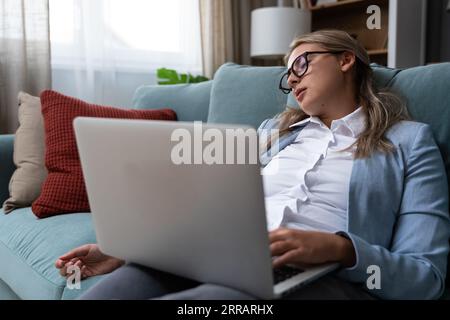 Junge Geschäftsfrau schlief auf dem Sofa ein, während sie am Laptop arbeitete. Überarbeitete, müde Frau, die es satt hat, vom Home Office aus am Computer zu arbeiten und mit mir geschlafen hat Stockfoto