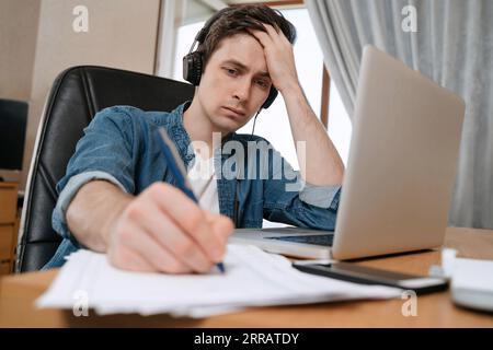 Müder junger Mann, der im Home Office an einem neuen Projekt arbeitet, frustrierter Kerl, der an seinem Schreibtisch sitzt und auf Papieren schreibt, über schwierige Aufgaben nachdenkt. Langweiliger Student, der von zu Hause aus studiert, zu Hause arbeitet. E-Learning Stockfoto