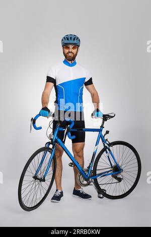 Glücklicher männlicher Radfahrer im Fahrradkit, der mit dem Fahrrad im leichten Studio steht. Vorderansicht eines bärtigen Radfahrers in blauem Jersey, der vor der Kamera lächelt, isoliert auf grauem Hintergrund. Konzept des Radsports, Aktivität. Stockfoto
