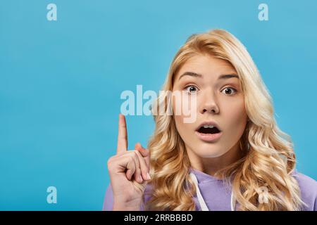 Porträt eines verblüfften blonden Teenagers mit offenem Mund, das Ideengeste auf Blau zeigt Stockfoto