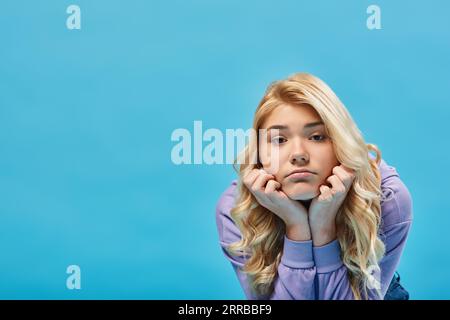 Frustriertes blondes Teenager-Mädchen im Hoodie, das die Hände nahe am Gesicht hält und die Kamera auf Blau anschaut Stockfoto