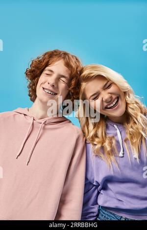 Fröhliche Teenager-Freunde in stylischen Hoodies lachen mit geschlossenen Augen bei blauen, sorglosen Teenagern Stockfoto