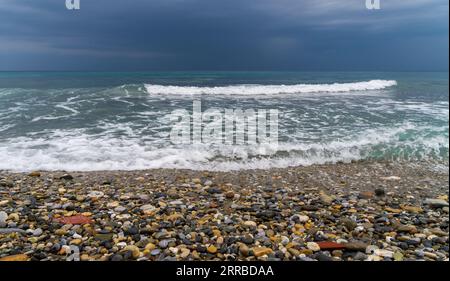 Dynamische Küstenlandschaft: Kleine, mehrfarbige Steine, die bei stürmischem Wetter mit schaumigen Wellen überflutet werden Stockfoto