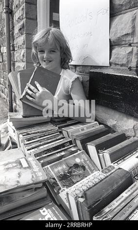 1983, Historic, ein junges Mädchen, das gebrauchte Bücher von einem Stand vor einem Viilage-Schreibwarenladen in Yorkshire, England, Großbritannien verkauft, um Geld für weihnachtsbeleuchtung in ihrem Dorf zu sammeln. Stockfoto