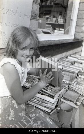 1983, Historic, ein junges Mädchen, das gebrauchte Bücher von einem Stand vor einem Viilage-Schreibwarenladen in Yorkshire, England, Großbritannien verkauft, um Geld für weihnachtsbeleuchtung in ihrem Dorf zu sammeln. Stockfoto