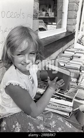 1983, Historic, ein junges Mädchen, das gebrauchte Bücher von einem Stand vor einem Viilage-Schreibwarenladen in Yorkshire, England, Großbritannien verkauft, um Geld für weihnachtsbeleuchtung in ihrem Dorf zu sammeln. Stockfoto