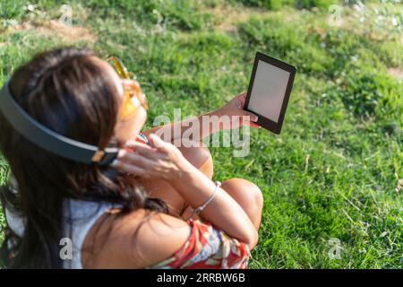 Frau mit Kopfhörern, die ein E-Book mit leerem Bildschirm hält, während sie im Park auf dem Rasen sitzt. Technologiekonzept. Stockfoto