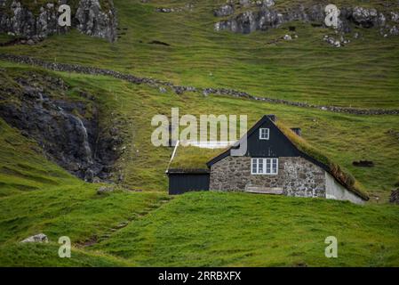 Ein Bauernhaus liegt auf einem Felsvorsprung mit einem steilen Berghang hinter Saksun auf der Insel Vagar auf den Färöern. Stockfoto