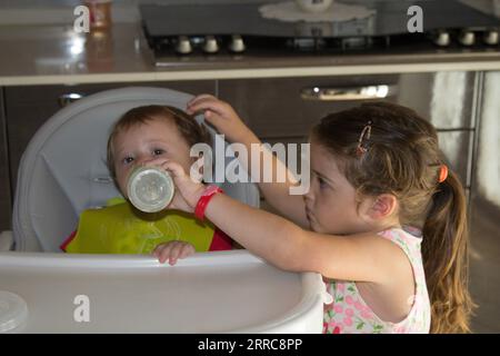 Bild eines entzückenden kleinen Mädchens, das seinem jüngeren Bruder im Hochstuhl mit einer Flasche Milch gibt. Brüderliche Liebe Stockfoto