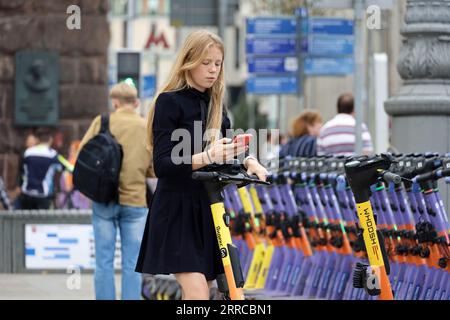 Mädchen mit Smartphone auf einer Straße mit Elektroroller auf dem Parkplatz. Transport und Reisen in der Stadt Stockfoto