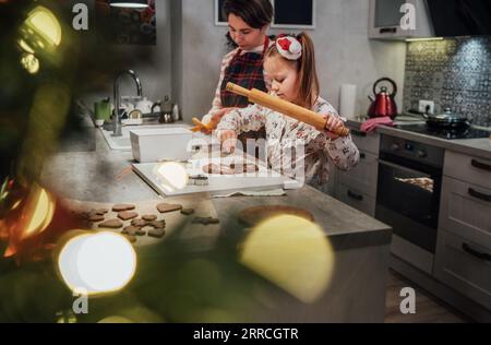 Niedliches kleines Mädchen mit Mutter, das selbstgemachte WeihnachtsLebkuchenkekse mit rollierender Nadel zusammen in der Hausküche macht. Glückliche Familienferien vorbereiten Stockfoto