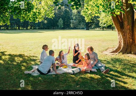 Große Familie, die am Wochenende auf einer Picknickdecke im Stadtpark auf Linden Treeb am Sonntagstag sitzt. Sie plaudern und essen gekochten Mais und Wassermelo Stockfoto