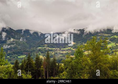 Sturmwolken Rollen über einen Berghang von Waldbäumen mit einem Dorf in der Ferne Stockfoto