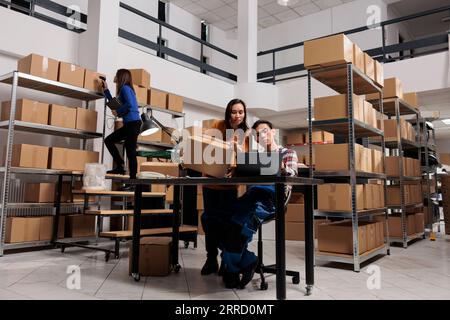 Postdienstmitarbeiter, die die Paketrechnung auf dem Laptop prüfen, bevor sie im Lagerraum arbeiten. Mitarbeiter im Lagerhaus, die Informationen zur Zollabfertigung auf dem Computer analysieren Stockfoto