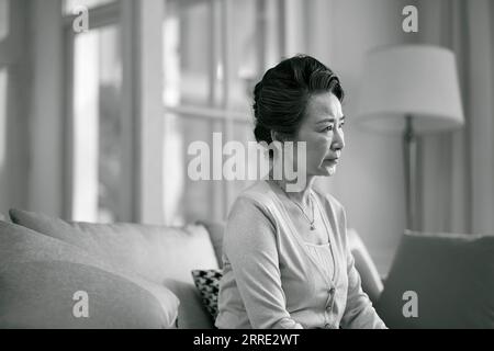 Seitenansicht der traurigen älteren asiatischen Frau, die allein auf der Couch im Wohnzimmer zu Hause sitzt, schwarz-weiß Stockfoto