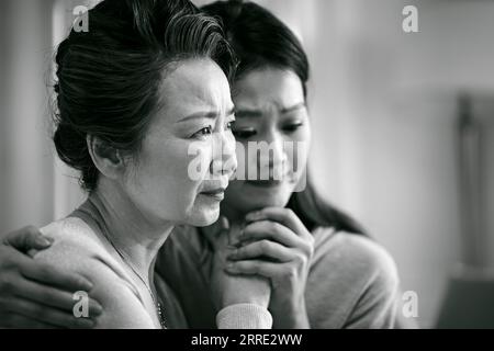 Traurige asiatische Seniormutter und erwachsene Tochter, die Hände hält, schwarz-weiß Stockfoto