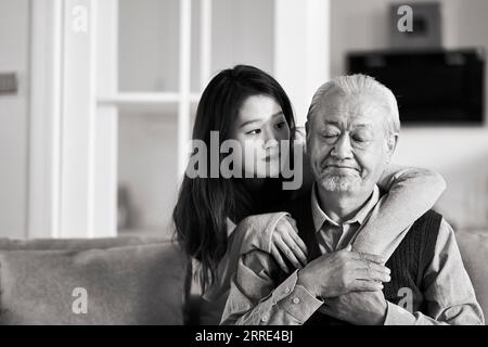 Trauriger älterer asiatischer Vater, der auf der Couch im Wohnzimmer zu Hause sitzt und von einer erwachsenen Tochter getröstet wird, schwarz-weiß Stockfoto